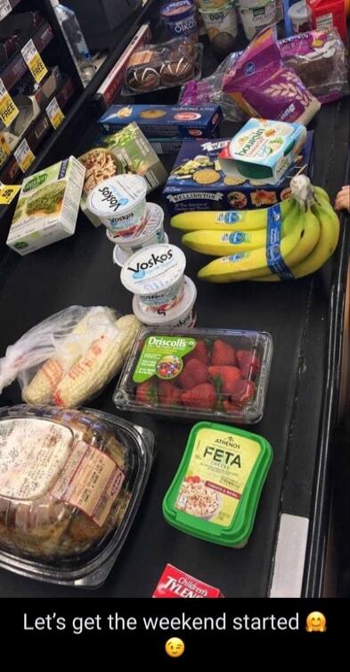 Let's get the weekend started. The image displays a variety of groceries on a checkout conveyor belt, including bananas, strawberries, corn on the cob, several containers of yogurt (Voskos, Oikos), feta cheese, crackers, and other packaged food items. A small box of Children's Tylenol is also visible.