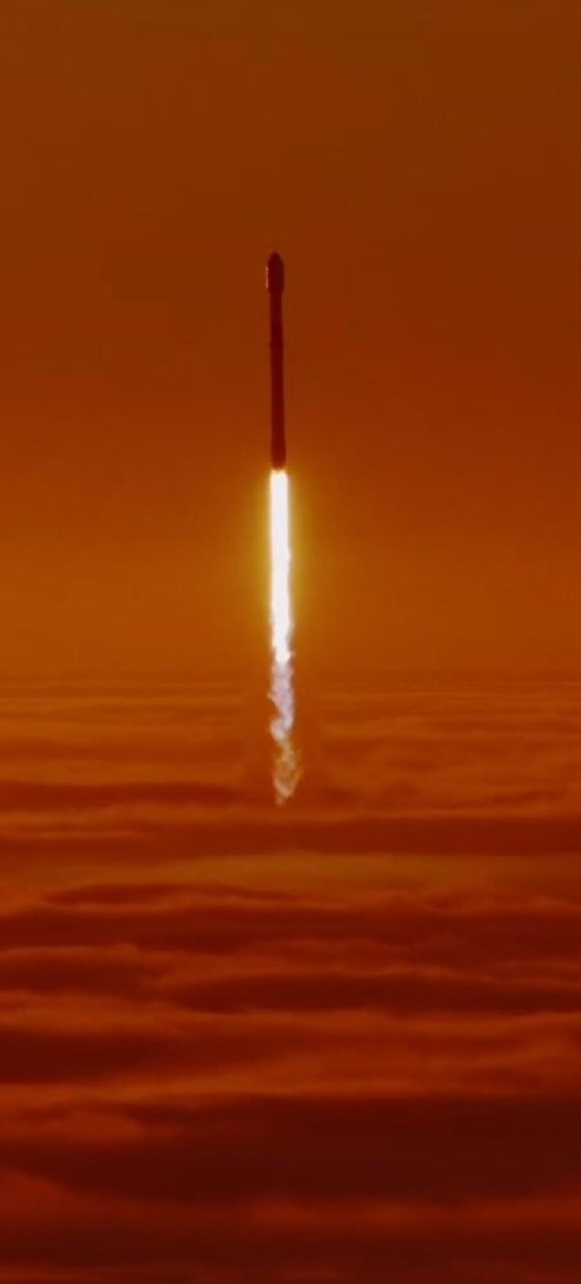 A rocket launches vertically into the sky, leaving a bright fiery plume as it rises above orange sunset clouds.
