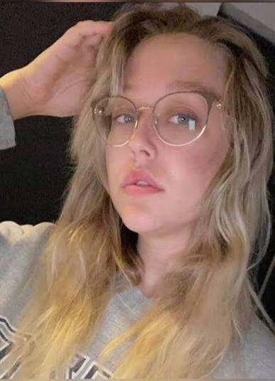 A young woman with blonde hair and glasses, wearing a grey sweatshirt, looking directly at the camera.