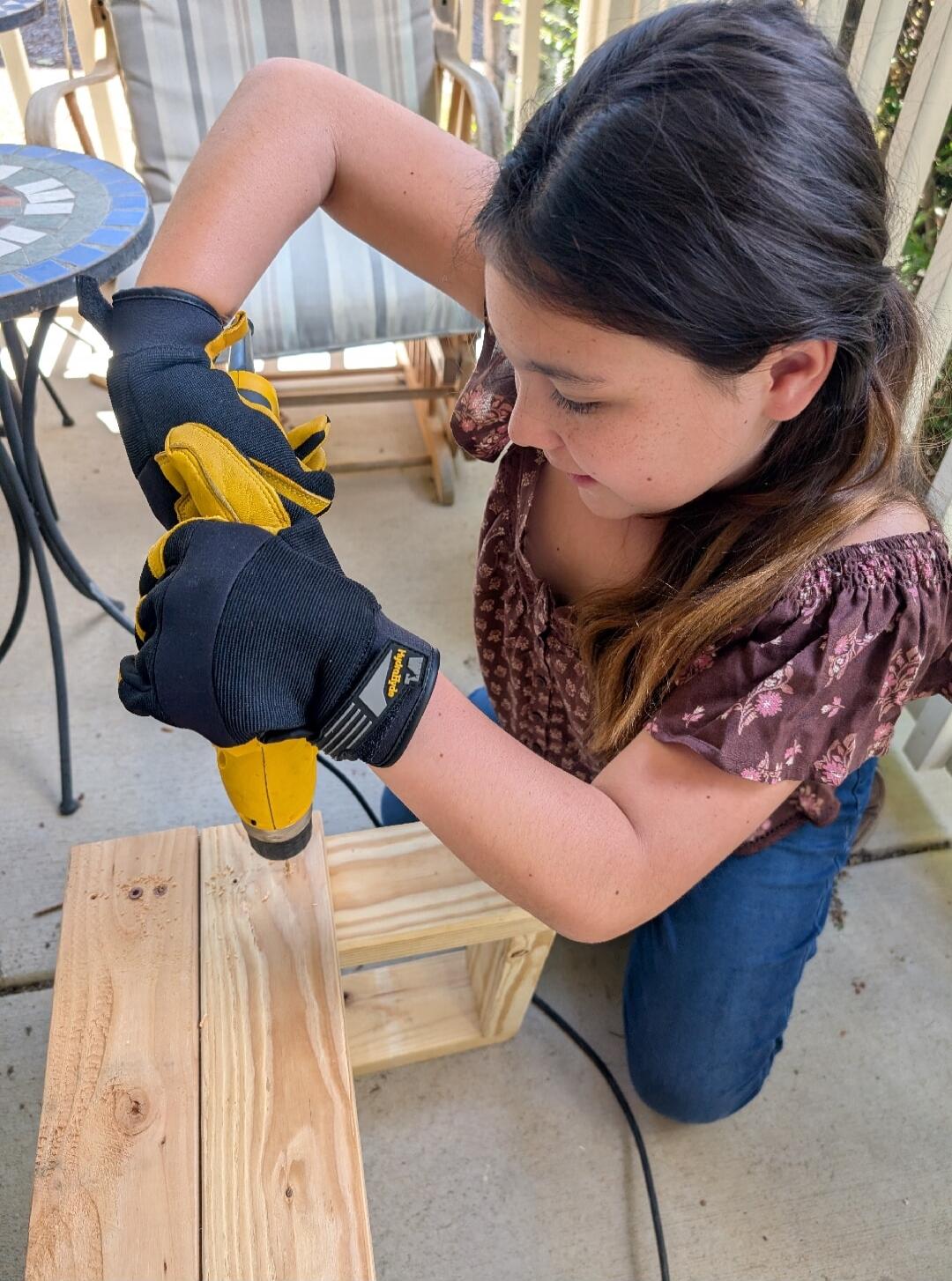 A young girl wearing black and yellow work gloves is using a yellow power drill to make a hole in a wooden plank. She is kneeling outdoors on a concrete surface, focused on her task. In the background, there is a patio chair and a small table with a mosaic top.