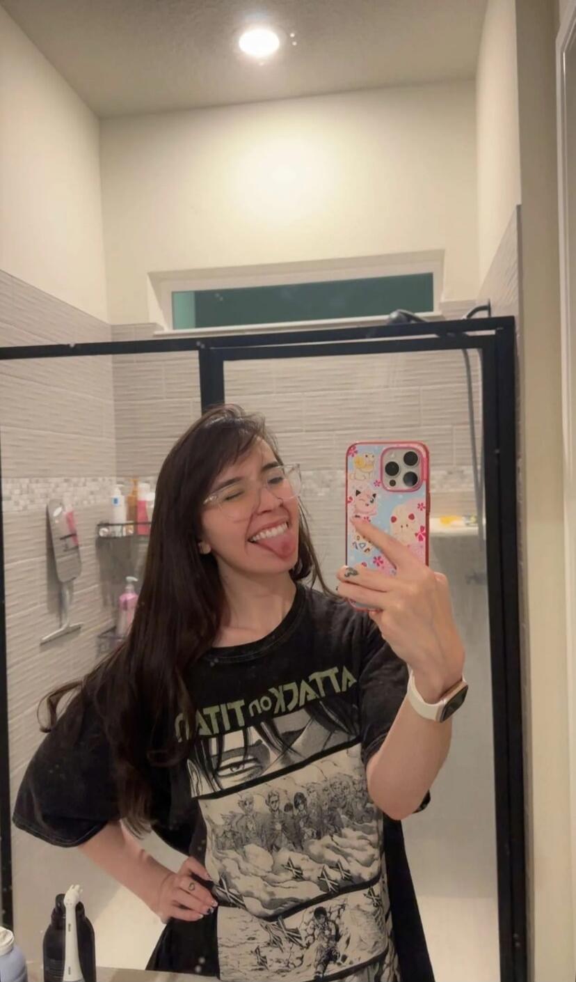 A young woman takes a mirror selfie in a bathroom, smiling and playfully sticking out her tongue. She is wearing glasses and a black t-shirt with an 'Attack on Titan' anime print. Her phone case has a cute, colorful design.