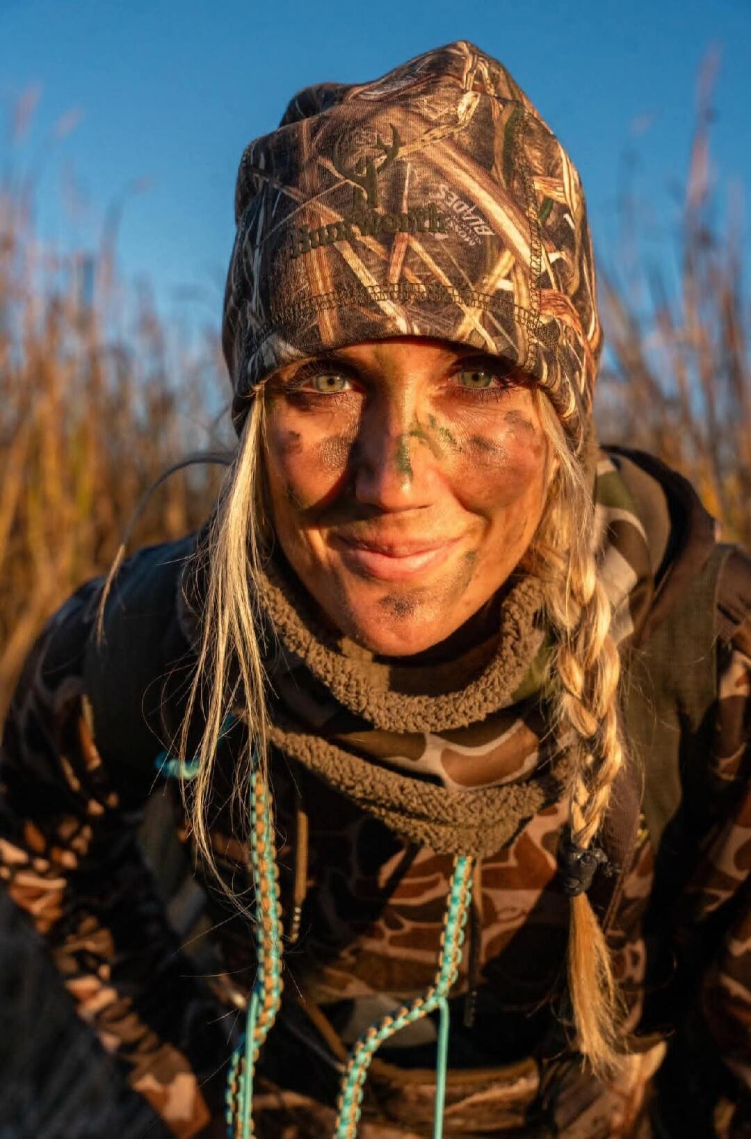 A woman in camouflage clothing and face paint smiles at the camera. Her beanie has 'MOSS BLADES' written on it.