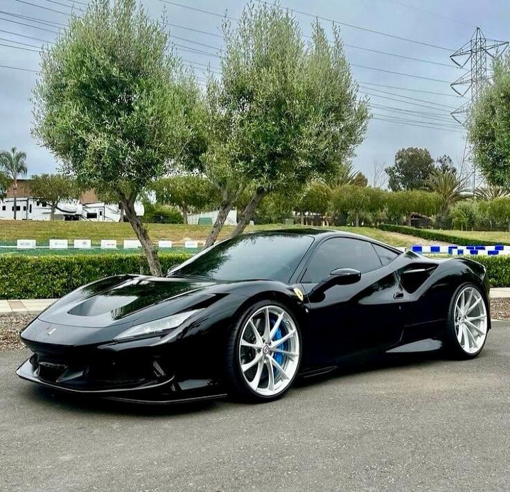A black Ferrari sports car is parked on an asphalt surface outdoors, with green trees and bushes in the background under a cloudy sky. The car features silver multi-spoke wheels with blue brake calipers.