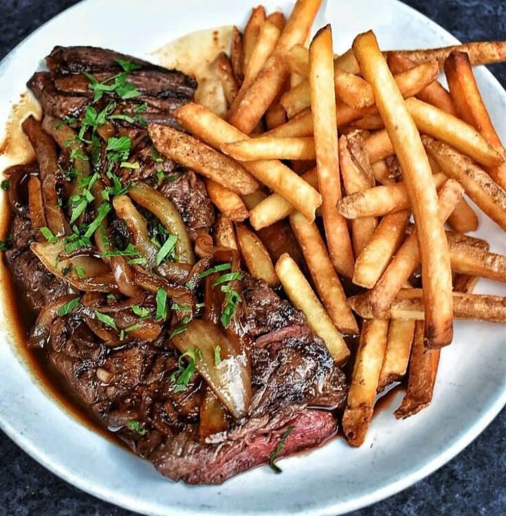 Cooked beef steak with onions, garnished with herbs, served with a pile of French fries.