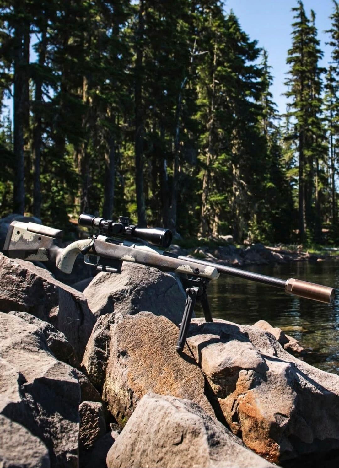 A sniper rifle with a scope and bipod rests on rocks near a body of water, with a forest in the background.