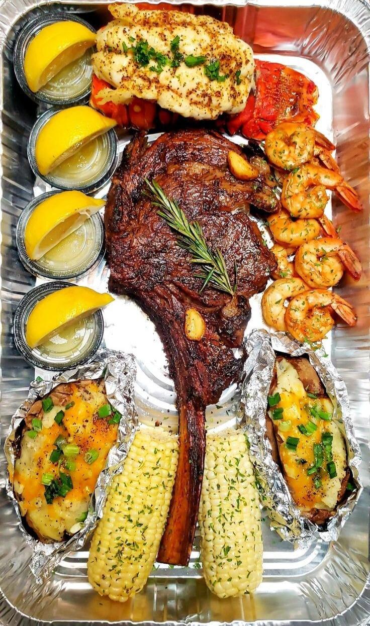 A delicious seafood and steak platter with lobster tail, grilled shrimp, a large tomahawk steak, corn on the cob, and baked potatoes, garnished with lemon wedges and fresh herbs.