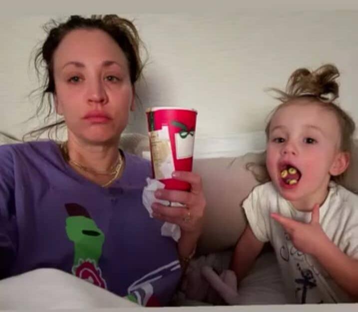 A tired woman with a messy bun holds a red cup and a tissue, looking exhausted. Next to her, a young child with cereal in their mouth points, looking mischievous.