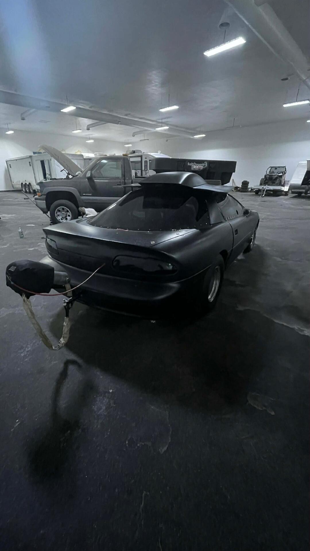 A dark coupe on a trailer in a large indoor workshop. The car appears to be connected to a tow hitch, with its trunk and rear area visible. Several other vehicles and workshop equipment are in the background.