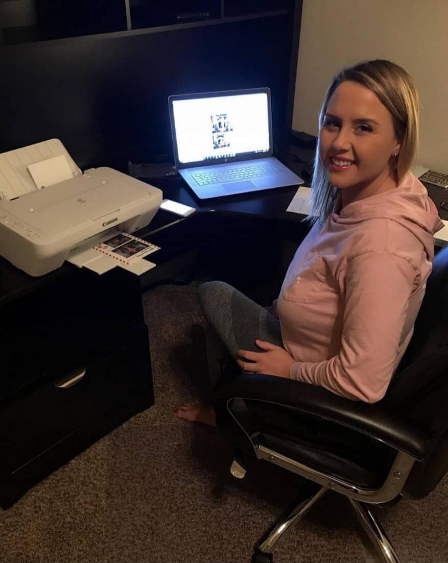 A woman is sitting at a desk with a printer on the left and a laptop open in front of her. She is smiling and wearing a light pink hoodie.
