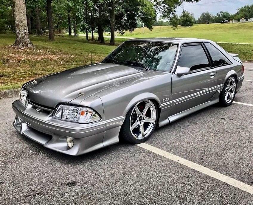 Silver modified Ford Mustang with 5.0 badge, large chrome wheels, front lip, and aftermarket body kit parked in a parking lot.