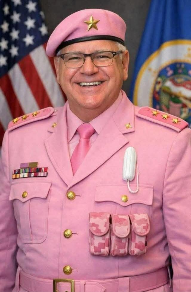 A man in a pink military-style uniform with gold buttons, ribbons, and pink pouches, wearing a pink beret with a star, smiling. American flag and another flag visible in the background.
