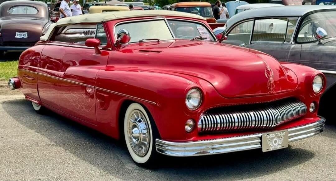 A vibrant red classic convertible car, possibly a Mercury, with custom pinstriping and whitewall tires, is parked outdoors. The front license plate reads 'White Wall Garage'. Other classic cars and people are visible in the background.