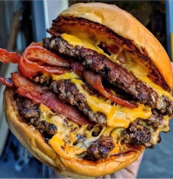 A heavily loaded bacon cheeseburger with multiple beef patties and melted cheese, topped with crispy bacon slices inside a soft bun.