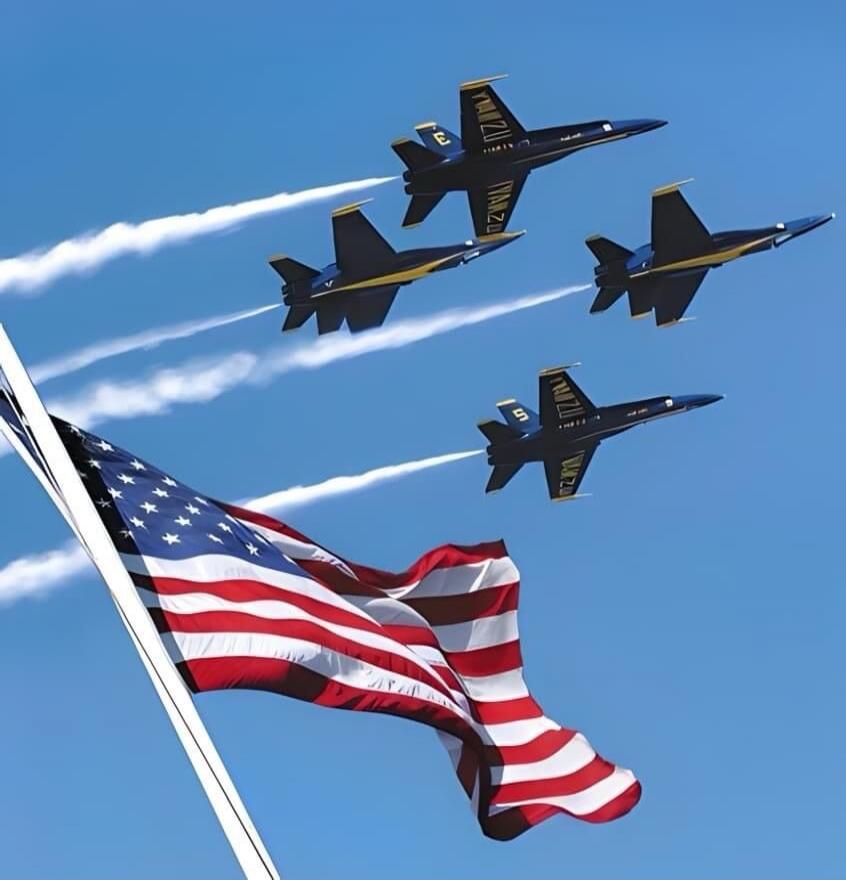 Four blue and yellow fighter jets, likely the Blue Angels, fly in formation, leaving white smoke trails across a clear blue sky. An American flag waves prominently in the foreground. The jets have 'NAVY' written on their wings and numbers like '3' and '5'.