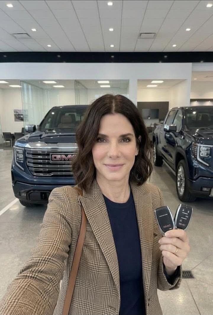 A woman, identified as Sandra Bullock, smiles while holding two car keys in a car dealership. Two blue GMC pickup trucks are visible in the background. The GMC logo is clearly visible on the front grille of the trucks.
