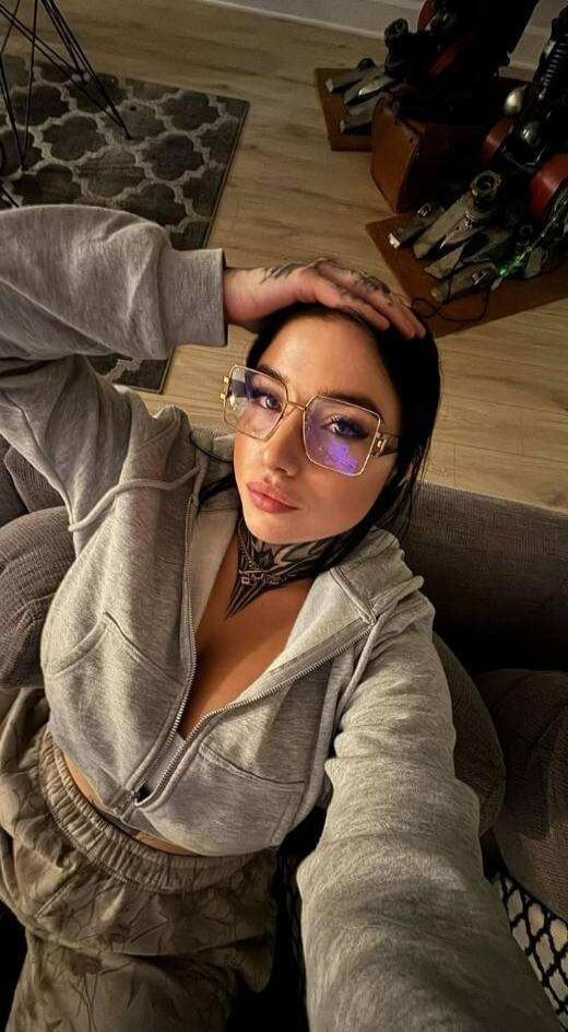 A woman wearing large glasses and a gray zip-up hoodie sits on a couch, with her hand resting on her head. She has a bold neck tattoo visible.