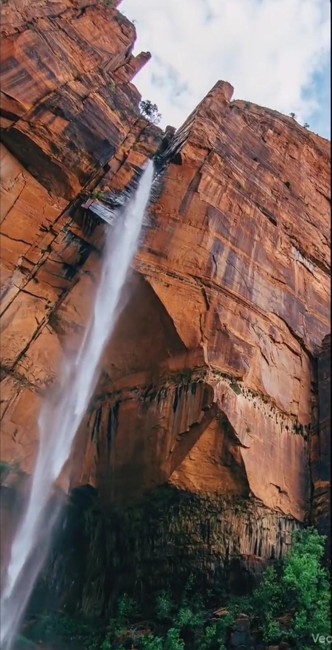 A tall waterfall streaming down a red rock cliff with lush green vegetation at the base.