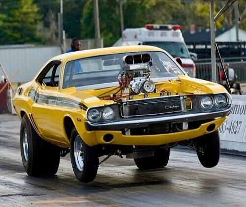 A yellow classic Dodge Challenger drag racing car with a large supercharger performs a wheelie on a drag strip. The front wheels are lifted off the ground as it accelerates. In the background, there's a person and an ambulance. Partial text 'mot 83-4627' is visible on a barrier.