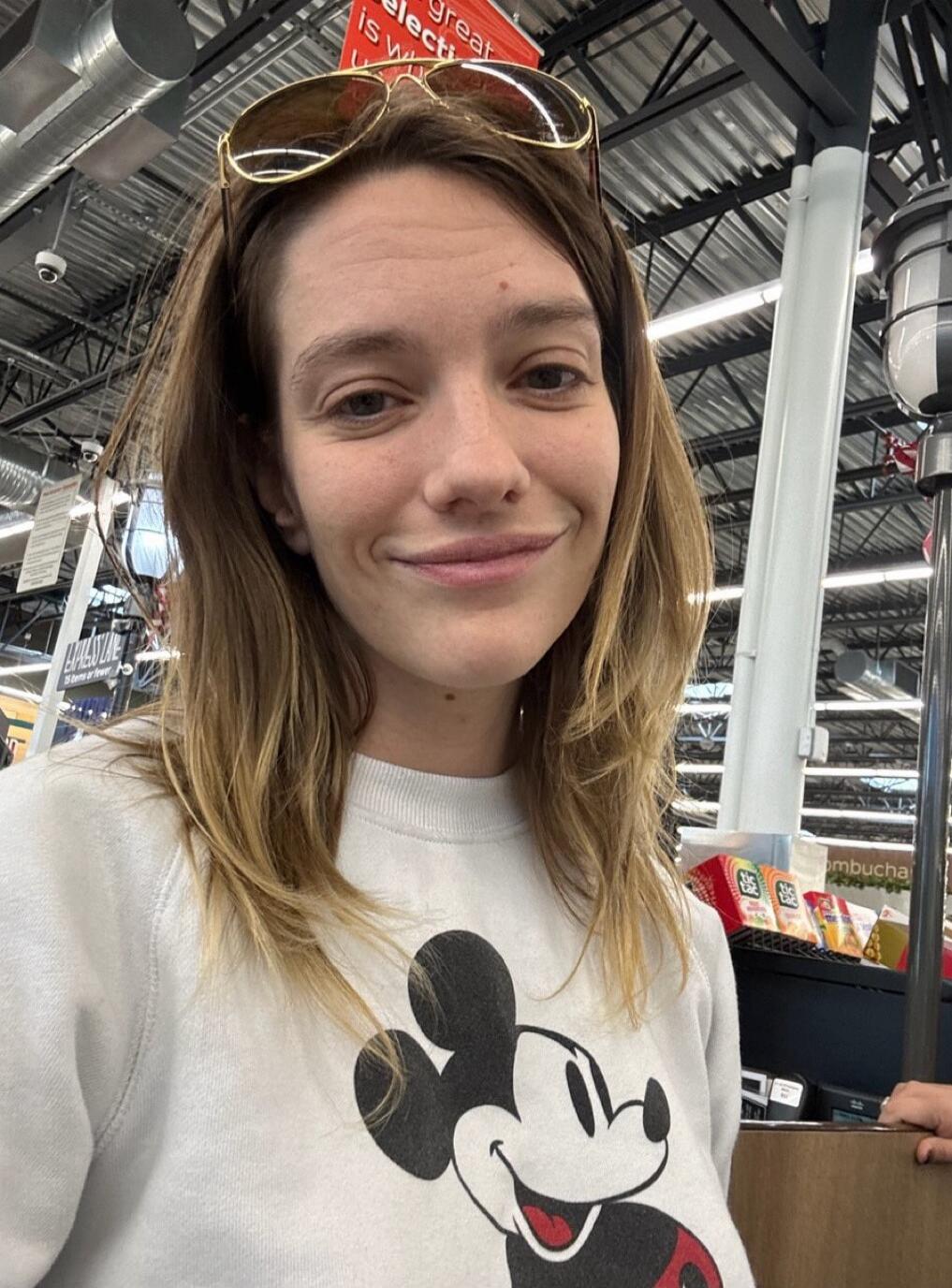 A young woman wearing a white sweatshirt featuring a Mickey Mouse graphic, sunglasses perched on her head, standing in a store with shelves and lighting visible in the background.