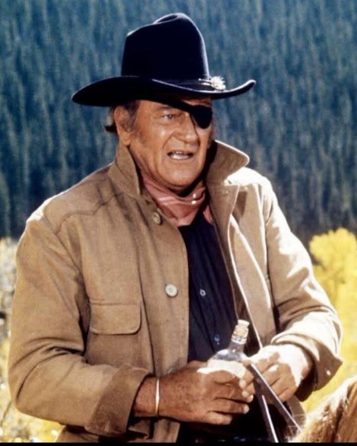 A man in a cowboy hat and an eye patch, wearing a jacket and a bandana, holding a bottle and a glass. He has a determined expression.