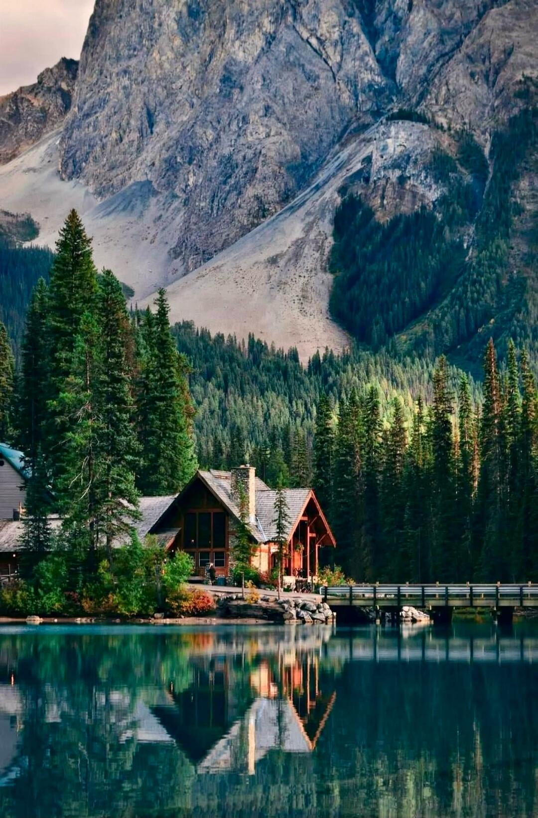 A cozy wooden cabin by a serene lake, surrounded by dense pine forests and towering mountain peaks in the background. The calm water perfectly reflects the scene.