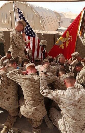 A group of soldiers in desert camouflage kneel and bow their heads in a moment of prayer or solemnity around a flag. American flag and Marine Corps flag visible in the background.