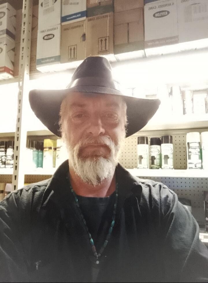 A man with a white beard and a dark hat is looking directly at the camera. He is wearing a dark shirt and a beaded necklace. The background shows shelves with boxes and spray cans, possibly in a store or workshop.