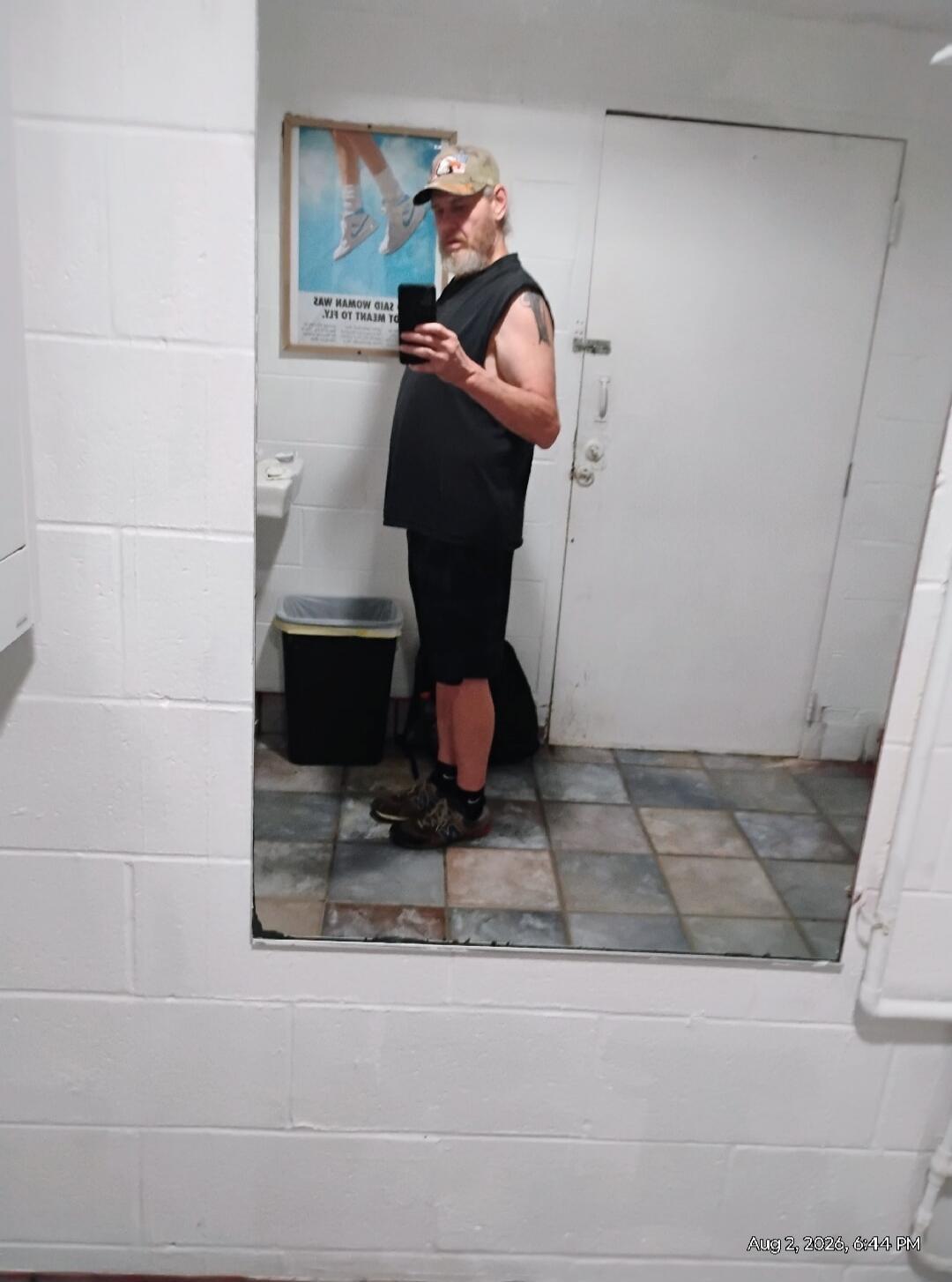 A man in a cap, black sleeveless shirt, and shorts takes a mirror selfie in a restroom. A poster with mirrored text is visible in the background, along with a sink and a trash can. The date and time 'Aug 2, 2026, 6:44 PM' are visible in the bottom right corner. The mirrored text on the poster appears to say 'SAID WOMAN WAS' and 'TO MEANT TO FLY'.