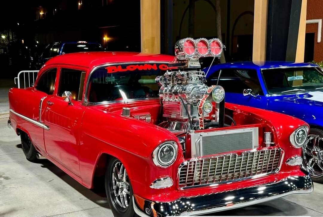 Red classic car with a prominent blower engine on display at a car show. chrome details, polished bumper, and another colorful car in the background.