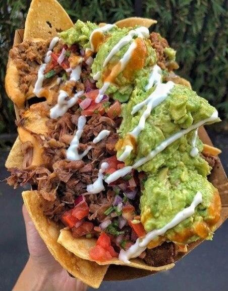 A close-up shot of a large serving of loaded nachos, featuring crispy tortilla chips topped with generous amounts of shredded meat, fresh guacamole, pico de gallo, and drizzled with white and orange sauces. The dish is held by a hand, suggesting it's a street food or casual dining item.