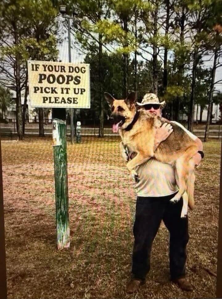 IF YOUR DOG POOPS PICK IT UP PLEASE. A man is seen carrying a large German Shepherd dog in his arms, standing next to a sign that reads 'IF YOUR DOG POOPS PICK IT UP PLEASE'. The man is wearing a hat and sunglasses, and the dog looks happy with its tongue out.