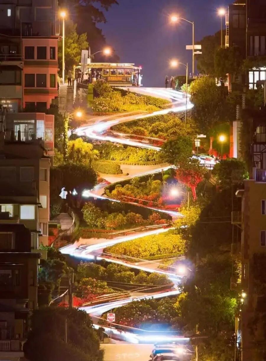 Long exposure photograph of Lombard Street in San Francisco at night, showing the famous winding road with car light trails and a cable car at the top. A 'STOP' sign is partially visible at the bottom.