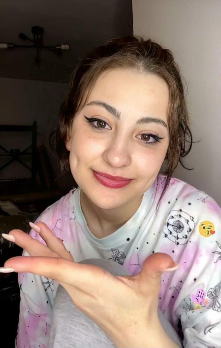 A young woman with dark hair and winged eyeliner smiles at the camera, showing her hand with long nails. She is wearing a tie-dye shirt with a winking kiss emoji.