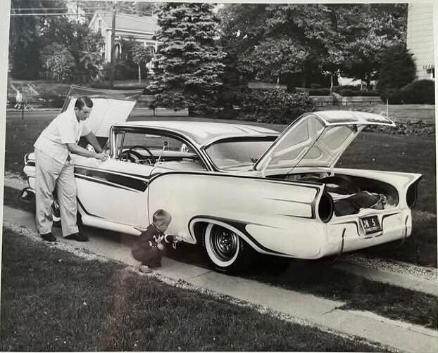 A man and a child standing by a vintage car with the hood and trunk open.
