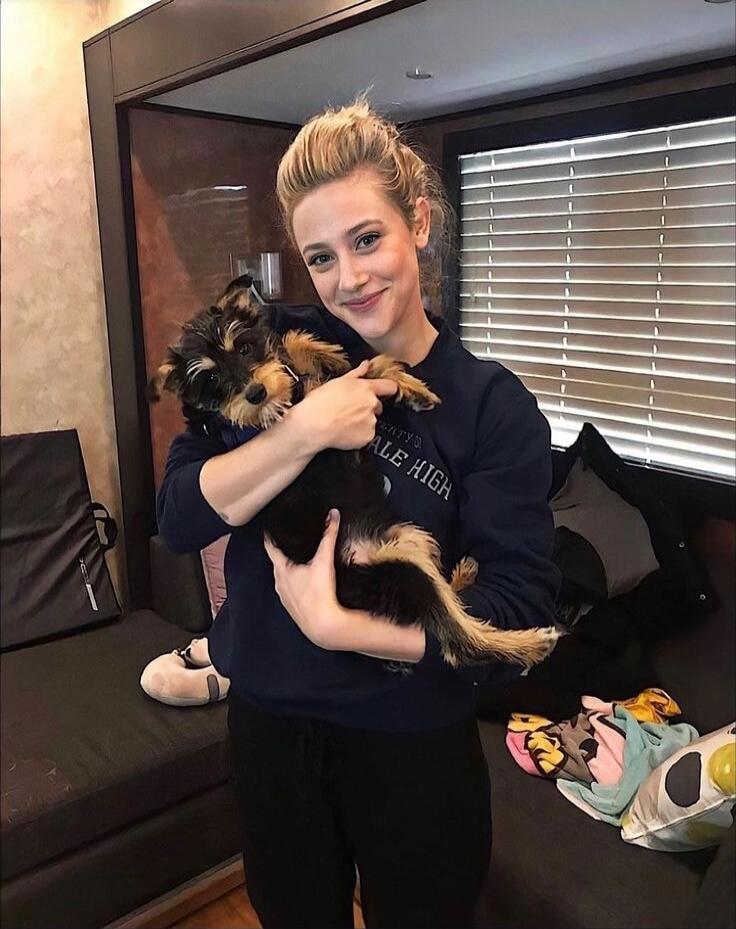 A young woman with blonde hair tied up, wearing a dark blue sweatshirt with 'RIVERDALE HIGH' visible, is smiling and holding a small, fluffy puppy with black and tan fur. The puppy is looking towards the camera. The background shows an indoor setting with blinds on a window and a dark couch.