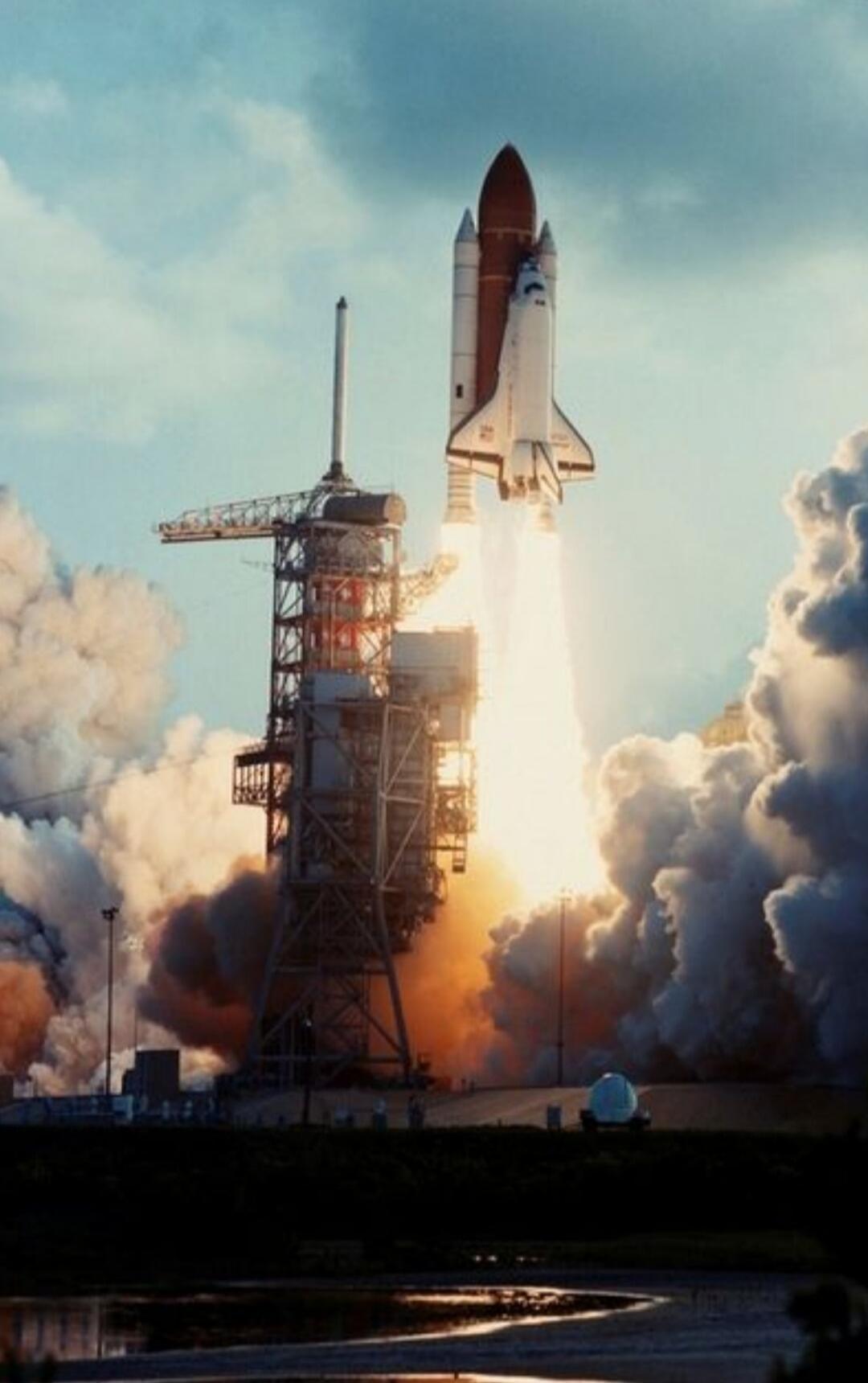 A space shuttle launching from a launchpad, with bright flames and smoke billowing from its engines. The shuttle is ascending into a partly cloudy sky, leaving a trail of exhaust.