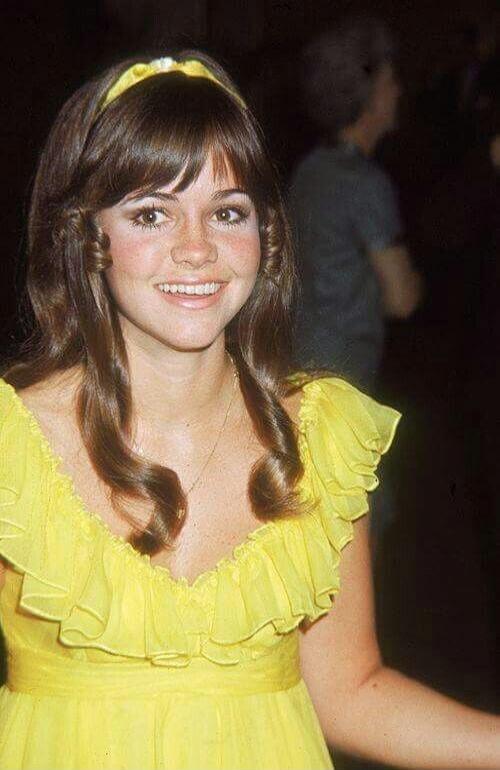 A young woman with long brown hair and bangs, wearing a yellow dress with ruffled sleeves and a yellow headband, smiles at the camera. She has freckles across her nose and cheeks.