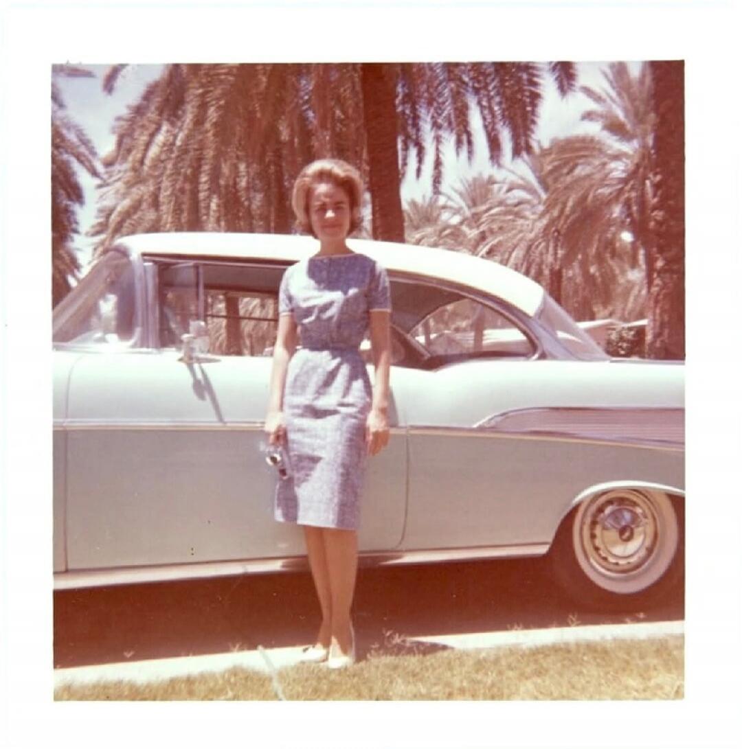 Photo of a woman standing beside a vintage car outdoors with palm trees in the background.