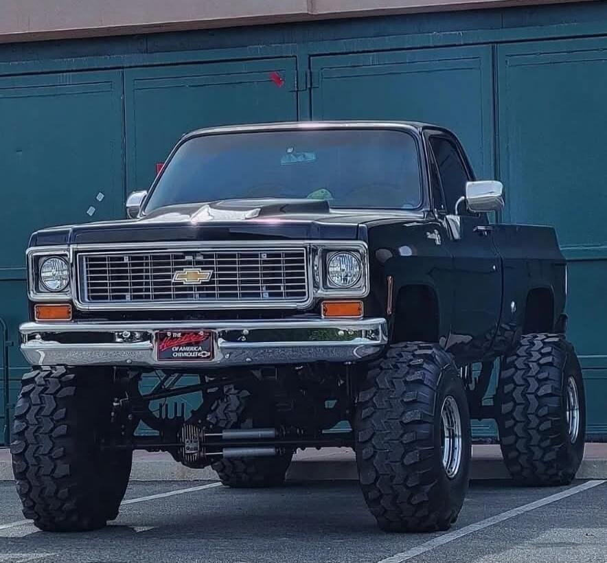 A dark green lifted Chevrolet pickup with oversized mud-terrain tires. The front grille features the Chevrolet bowtie emblem; chrome bumper stands out. The vehicle is parked in a lot in front of a blue garage door.
