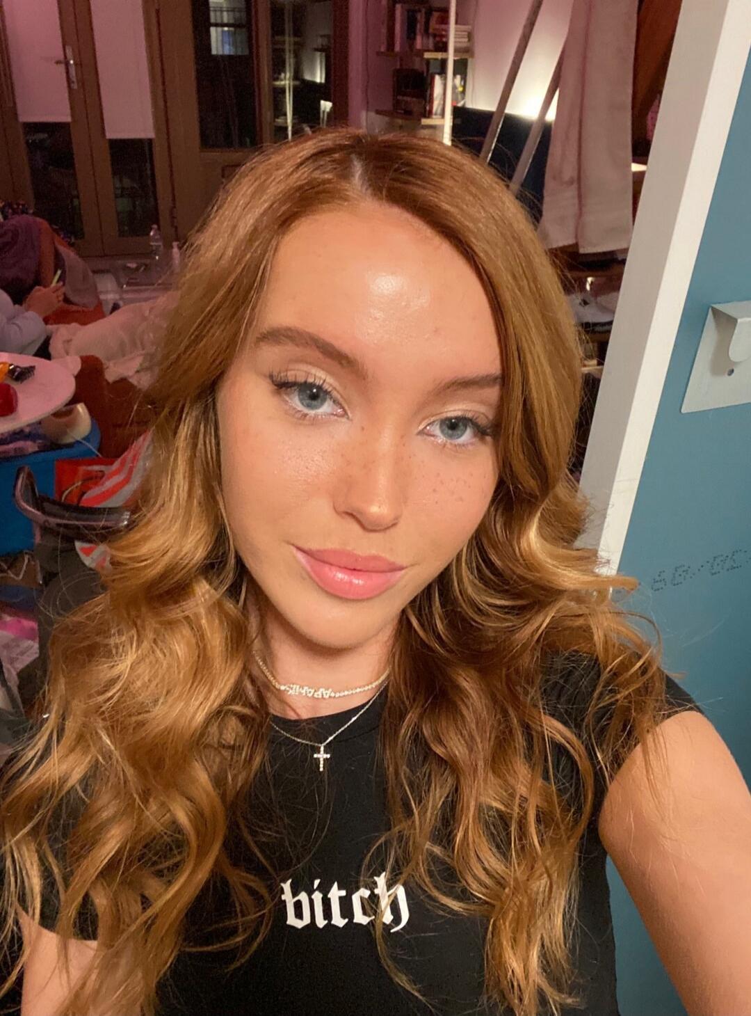 The woman's black t-shirt has the word 'bitch' printed on it. She is also wearing a necklace with the word 'APARTIS'. In the background, on a blue wall, there are faint markings 'SB-00-05'.
