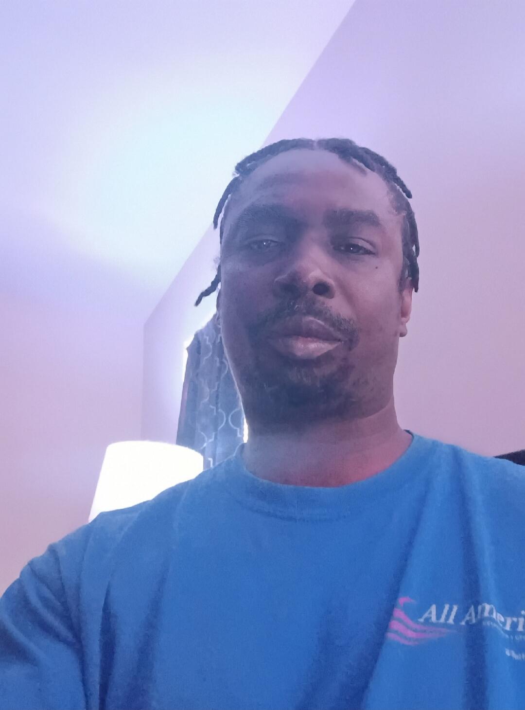 A man with braided hair wearing a blue t-shirt with 'All Ameri' visible on it, looking at the camera.