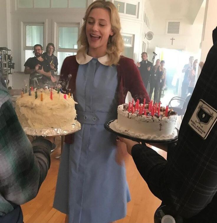 A woman smiling widely at two birthday cakes with lit candles, held by people in front of her. The scene appears to be on a film set, with crew members visible in the background. A partial sign 'RY' is seen, and a badge on a person's sleeve reads 'NOEL ASHMAN EQUESTRIAN'.