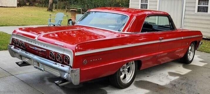 Red 1960s Chevrolet Impala coupe parked on a driveway, classic chrome bumper and styling.