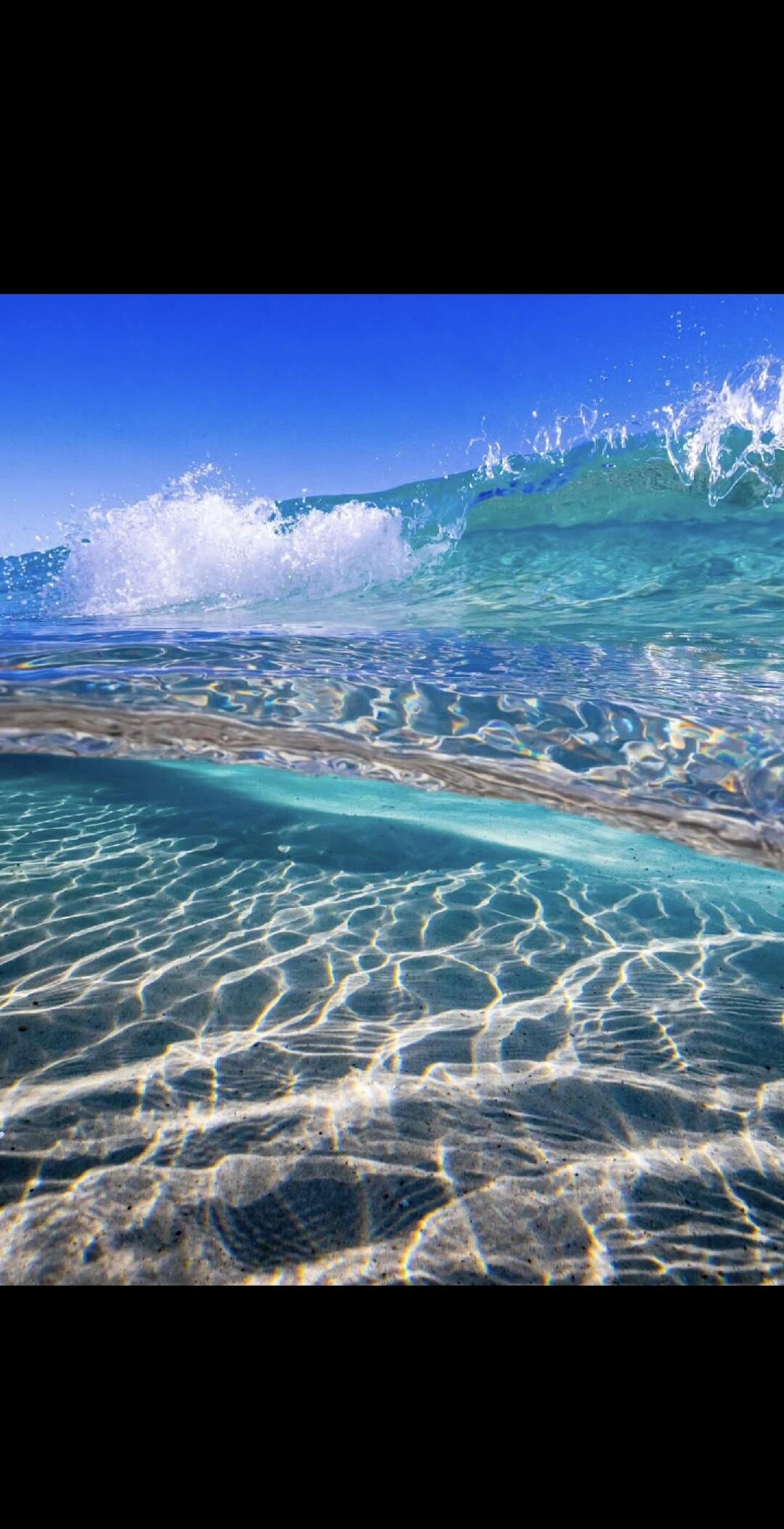 The image shows a clear blue sky above a breaking wave in the ocean. The water is transparent, revealing the sandy seabed with sunlight patterns. The perspective is from underwater, looking up towards the surface and the breaking wave.