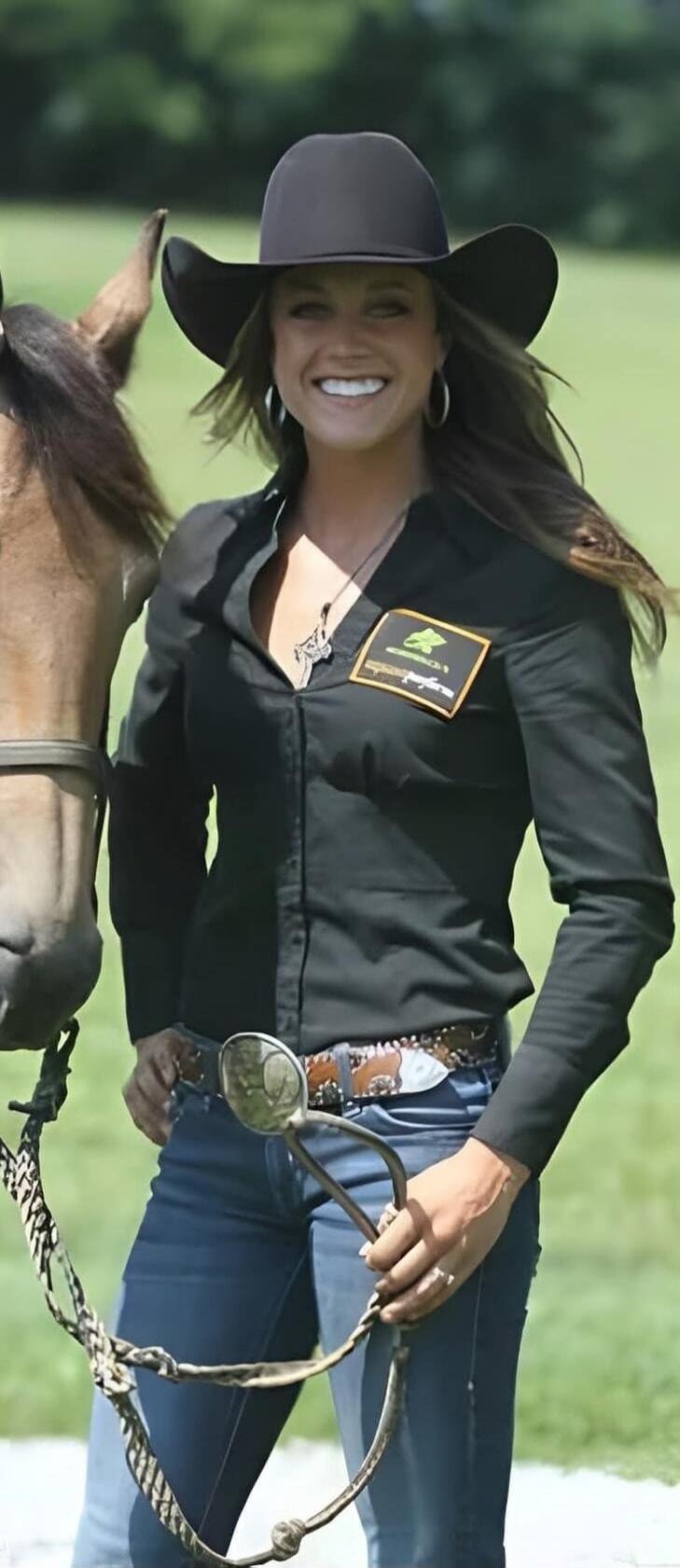 A smiling woman in a black cowboy hat, black long-sleeved shirt, blue jeans, and a large belt buckle stands next to a horse. She is holding a bridle or reins. There is a patch on her shirt with a green logo and some text that is difficult to read. The background is a green field.