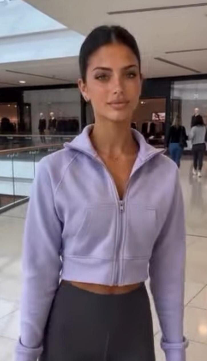 A young woman with dark hair and light eyes, wearing a light purple cropped zip-up jacket and dark leggings, stands in a modern indoor setting, possibly a shopping mall. She looks directly at the camera with a neutral expression. Other people and mannequins are visible in the blurred background.