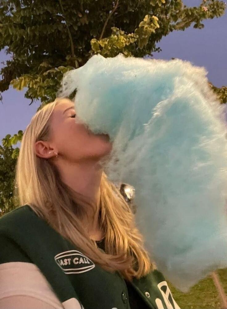 A woman in a green jacket is blowing a large cloud of blue cotton candy from her mouth.
