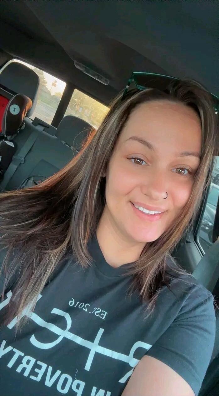 A woman is smiling in a car. She is wearing a black t-shirt with white text that reads 'IM POVERTY EROS.23' (text is mirrored in the image).