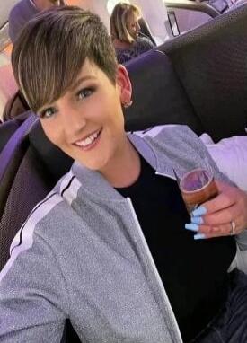 Smiling person with short hair wearing a grey jacket, holding a pink beverage in a clear cup.