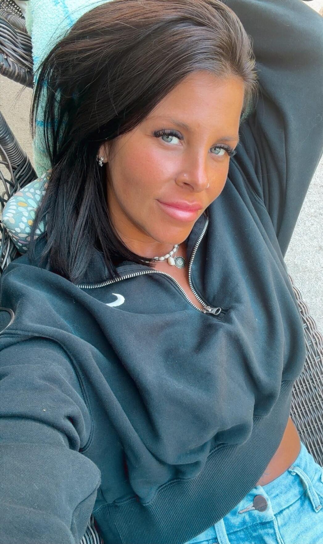 A young woman with dark hair and blue eyes, wearing a black half-zip sweatshirt and light blue jeans, is lying down and looking directly at the camera.
